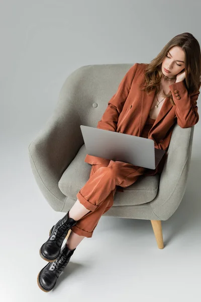 Overhead view of brunette young woman with long hair and necklace wearing trendy suit with blazer and pants and using laptop while sitting in armchair on grey background, freelancer — Stock Photo