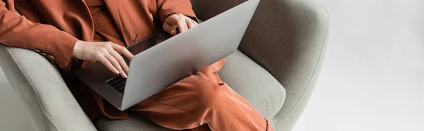 Vista superior de la mujer joven que usa traje de moda de terracota con chaqueta y pantalones usando el ordenador portátil mientras está sentado en cómodo sillón sobre fondo gris, freelancer, trabajo remoto, tiro recortado, pancarta - foto de stock