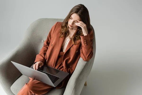 Tired young woman with long hair wearing terracotta trendy suit with blazer and pants and using laptop while sitting in comfortable armchair on grey background, freelancer, remote work — Stock Photo