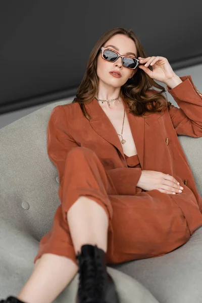 Stylische junge Frau mit langen Haaren trägt Terrakotta-Anzug mit Blazer und Hose und posiert mit trendiger Sonnenbrille, während sie in verschwommenen Stiefeln auf einem Sessel auf grauem Hintergrund sitzt, modisches Modell — Stockfoto