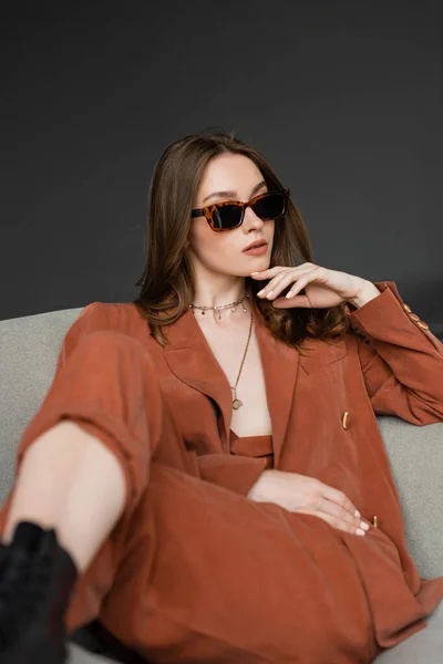 Stylische junge Frau mit langen Haaren trägt Terrakotta-Anzug mit Blazer und Hose und posiert mit trendiger Sonnenbrille, während sie im bequemen Sessel auf grauem Hintergrund sitzt, modisches Modell, schaut weg — Stockfoto