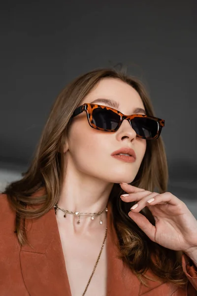 Portrait of stylish young woman with long hair wearing suit with terracotta blazer and posing in trendy sunglasses while looking away near blurred armchair on grey background, fashionable model — Stock Photo