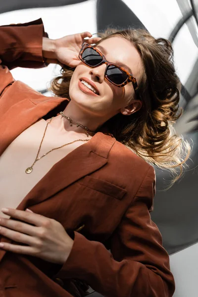Positive Frau mit langen Haaren in Terrakotta und trendigem Anzug mit Blazer und goldener Halskette, posiert mit Sonnenbrille auf abstrakt grauem Hintergrund, schönes Modell, Sonnenlicht — Stockfoto