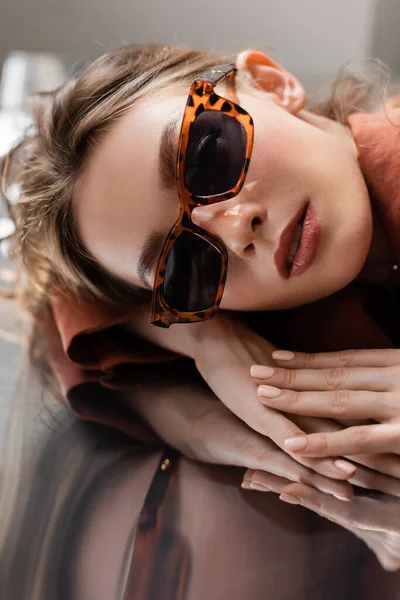 Jovem bonita com cabelo morena vestindo óculos de sol na moda e olhando para a câmera enquanto deitado sobre fundo espelhado cinza, modelo bonito, reflexão — Fotografia de Stock