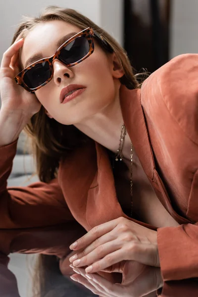 Junge Frau in Terrakotta und trendigem Anzug mit Blazer und goldener Halskette und posiert in modischer Sonnenbrille auf grau verspiegeltem Hintergrund, schönes Modell, Spiegelung, Blick in die Kamera — Stockfoto