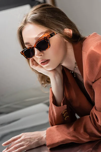 Mujer joven con traje de moda con chaqueta de terracota y collar de oro y posando en gafas de sol de moda sobre fondo gris espejo, hermoso modelo, reflexión, soñador - foto de stock
