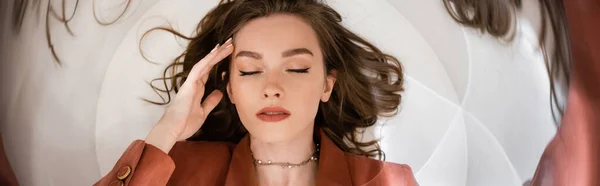 Circular shot, top view of young woman with brunette hair wearing blazer and golden necklace while lying and touching face on grey background, blurred, wide-angle, abstract composition, banner — Stock Photo