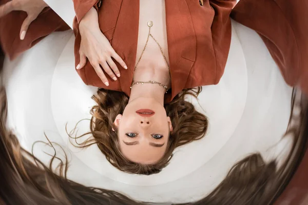 Circular shot, top view of young woman with brunette hair wearing blazer and golden necklace while lying and looking at camera on grey background, blurred, abstract composition, creative perspective — Stock Photo