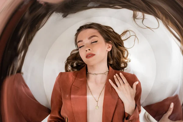 Circular shot of young woman with brunette hair and closed eyes wearing blazer and golden necklace while lying on grey background, blurred, wide-angle, distorted view, top view, creative perspective — Stock Photo