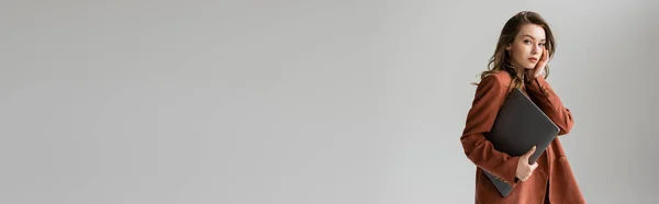 Mujer joven con cabello moreno y collar con traje de moda de terracota con chaqueta, mirando a la cámara y sosteniendo el ordenador portátil mientras está de pie sobre fondo gris, freelancer, trabajo remoto, pancarta - foto de stock
