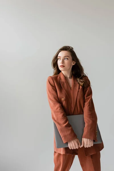 Young woman with brunette hair and necklace wearing terracotta and trendy suit with blazer and pants, looking away and holding laptop while standing on grey background, freelancer, remote work — Stock Photo