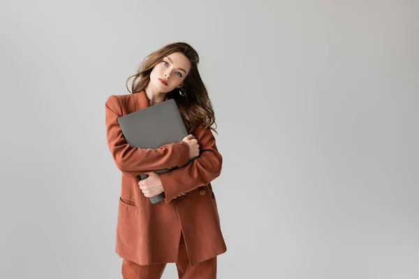 Pretty freelancer with brunette hair in terracotta and trendy suit with blazer and pants, holding laptop while standing on grey background, looking camera, young woman, remote work — Stock Photo