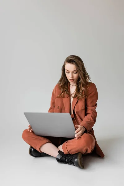 Brunette young woman in golden necklace sitting with crossed legs in terracotta and trendy suit, using laptop while working remotely on grey background, freelancer, digital nomad — Stock Photo