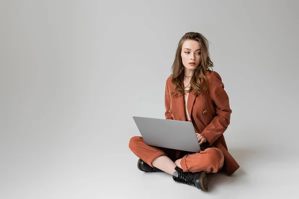 Brünette junge Frau in goldener Halskette sitzt mit überkreuzten Beinen in Terrakotta und trendigem Anzug, benutzt Laptop, während sie ferngesteuert auf grauem Hintergrund arbeitet, Freelancer, digitaler Nomade, schaut weg — Stockfoto