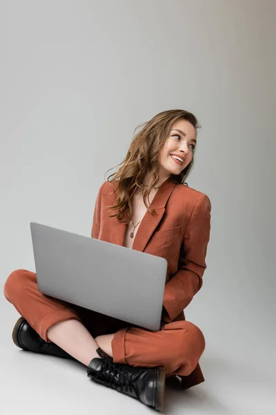 Cheerful young woman in golden necklace sitting with crossed legs in  trendy suit, using laptop, looking away while working remotely on grey background, freelancer, digital nomad — Stock Photo