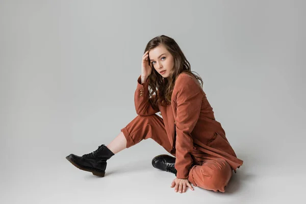 Full length of fashionable young woman with brunette and wavy hair sitting in trendy and oversize suit with blazer, pants and black boots, looking at camera on grey background, stylish model — Stock Photo