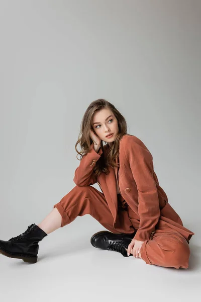 Full length of fashionable young woman with brunette and wavy hair sitting in trendy and oversize suit with blazer, pants and black boots, looking away on grey background, stylish model — Stock Photo