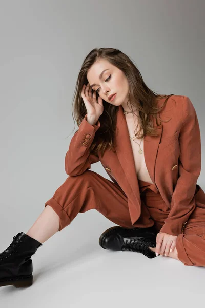 Full length of sensual and young woman with brunette and wavy hair sitting in trendy and oversize suit with blazer, pants and black boots, looking down on grey background, stylish model — Stock Photo