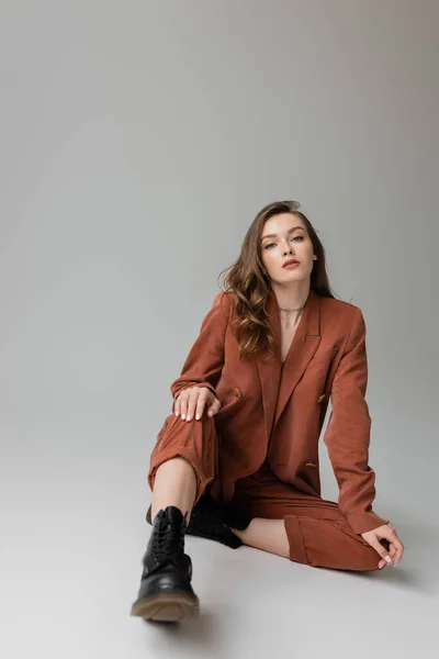 Full length of fashionable woman with brunette and wavy hair sitting in trendy and oversize suit with blazer, pants and black boots, looking at camera on grey background, young model — Stock Photo