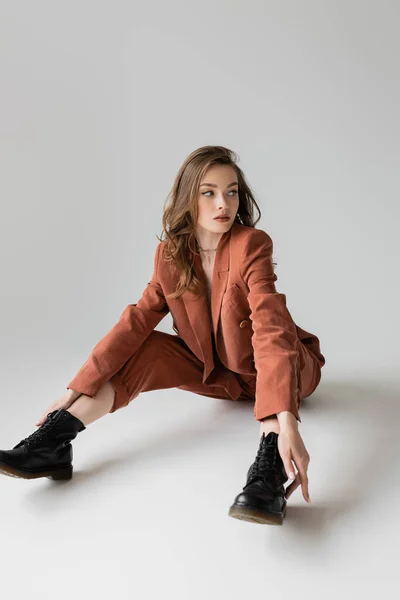 Full length of chic young woman with brunette and wavy hair sitting in trendy and oversize suit with blazer, pants and black boots, looking away on grey background, stylish model — Stock Photo