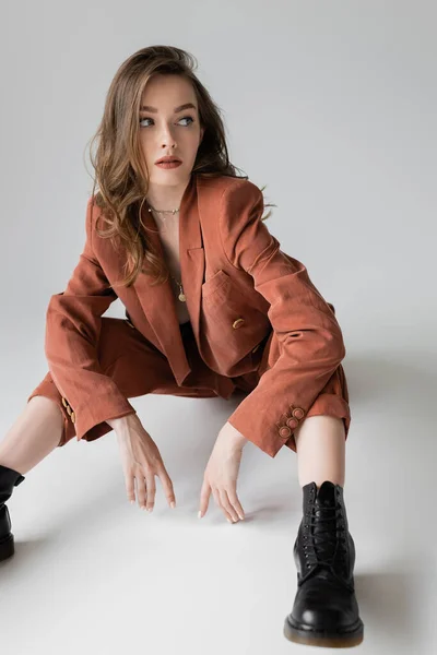 Chic jeune femme aux cheveux bruns et ondulés assis en costume tendance et oversize avec blazer, pantalon et bottes noires, regardant loin sur fond gris, modèle élégant, glamour — Photo de stock
