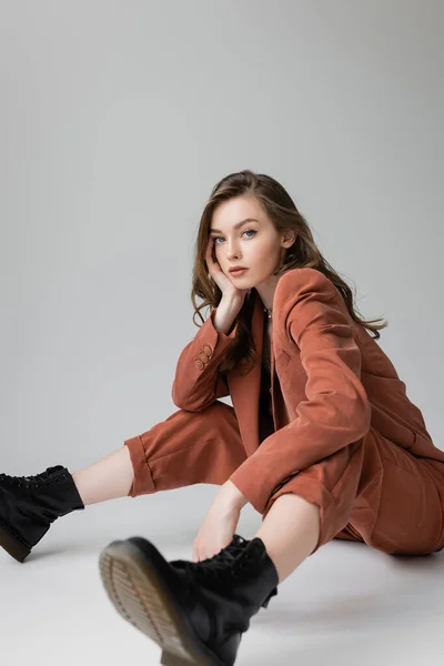Full length of fashionable model with brunette and wavy hair sitting in trendy and oversize suit with blazer, pants and black boots, looking at camera on grey background, stylish young woman — Stock Photo