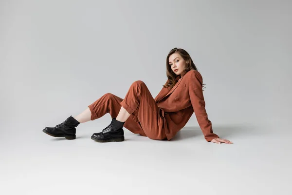 Full length of trendy model with brunette and wavy hair sitting in trendy and oversize suit with terracotta blazer, pants and black boots, looking at camera on grey background, stylish young woman — Stock Photo