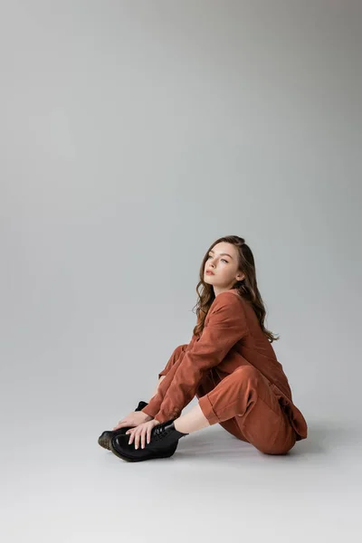 Full length of dreamy model with brunette and wavy hair sitting in trendy and oversize terracotta suit with blazer, pants and black boots, looking away on grey background, young woman, stylish pose — Stock Photo