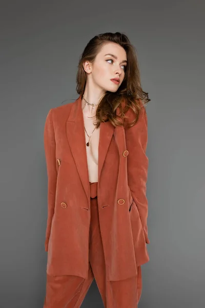 Young shirtless woman with brunette and wavy hair posing in trendy and terracotta suit with oversize blazer and golden necklace, looking away on grey background,  stylish pose, sexy model — Stock Photo