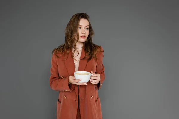 Mujer joven y sofisticada con cabello moreno que usa traje marrón y de moda con chaqueta y taza de café mientras mira hacia otro lado sobre fondo gris, equilibrio vida-trabajo - foto de stock