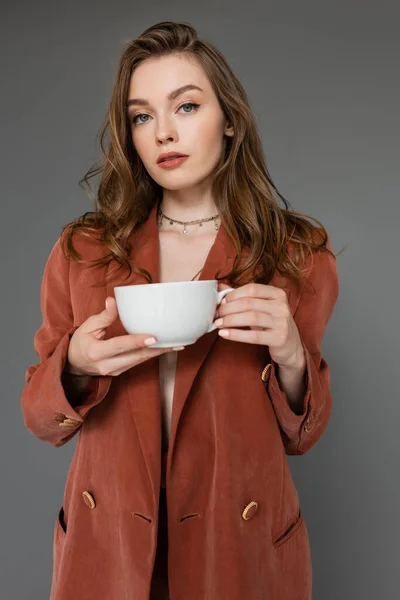 Sophisticated young woman with brunette hair wearing brown and trendy suit with blazer and holding cup of coffee while looking at camera on grey background, work-life balance — Stock Photo