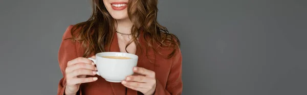 Vue recadrée d'une jeune femme heureuse aux cheveux bruns portant un costume marron et tendance avec un blazer et tenant une tasse de café sur fond gris, équilibre travail-vie privée, bannière — Photo de stock