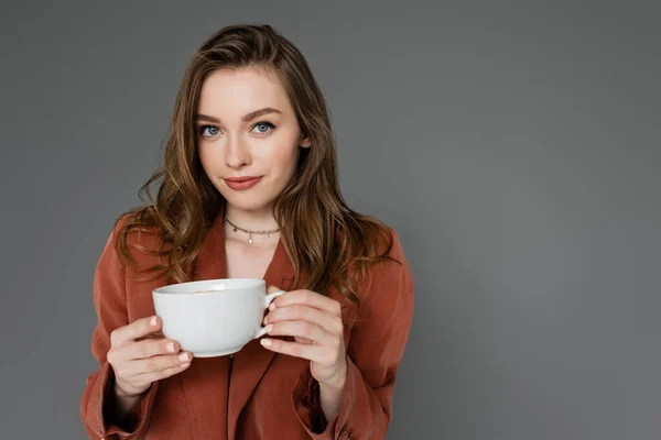 Attractive young woman with brunette hair wearing brown and trendy suit with blazer and holding cup of coffee while looking at camera on grey background, work-life balance — Stock Photo