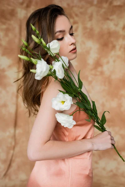 Portrait de brune et jeune femme en soie rose slip robe tenant des fleurs d'eustomie tout en se tenant debout sur fond beige marbré, sensualité, élégance, sophistication — Photo de stock