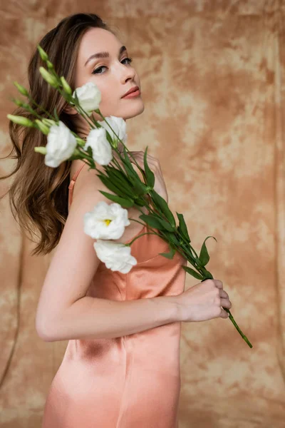 Retrato de elegante, morena y mujer joven en vestido de seda rosa deslizamiento sosteniendo flores eustoma mientras está de pie sobre fondo beige moteado, sensualidad, elegancia, sofisticación, mirando a la cámara - foto de stock