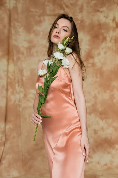 Morena e jovem mulher de seda rosa vestido deslizante segurando flores eustoma enquanto de pé e olhando para a câmera no fundo bege manchado, sensualidade, elegância, sofisticação — Fotografia de Stock