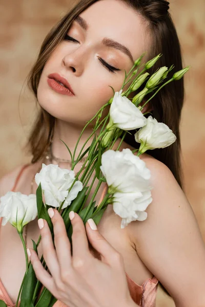 Retrato de mulher sonhadora, morena e jovem com olhos fechados segurando flores eustoma enquanto em pé e posando sobre fundo bege manchado, sensualidade, elegância, sofisticação — Stock Photo