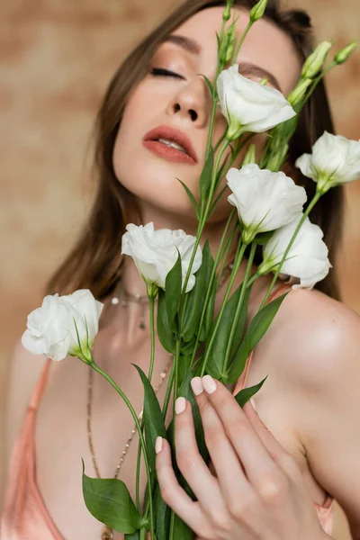 Portrait de femme sensible et jeune en soie rose glissante tenant des fleurs d'eustomie debout les yeux fermés sur fond beige marbré, sensualité, élégance, sophistication — Photo de stock