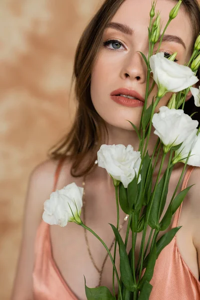 Portrait of sensual and young woman holding eustoma flowers while standing and looking at camera on mottled beige background, captivating beauty, elegance, sophistication — Stock Photo