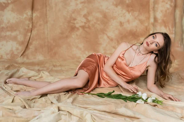 Longitud completa de descalzo y sensual mujer joven en vestido de seda rosa deslizamiento acostado en tela de lino cerca de flores blancas sobre fondo beige moteado, sofisticación, mirando a la cámara, eustoma - foto de stock