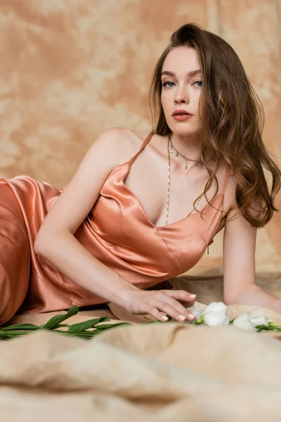Pretty young woman in pink silk slip dress lying on blurred linen fabric and looking at camera near white flowers on mottled beige background, sensuality, sophistication, elegance, eustoma — Stock Photo