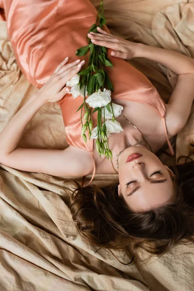Vista superior de la mujer joven en vestido de seda rosa deslizamiento acostado en tela de lino con los ojos cerrados y la celebración de flores blancas sobre fondo beige moteado, sensualidad, sofisticación, elegancia, eustoma - foto de stock