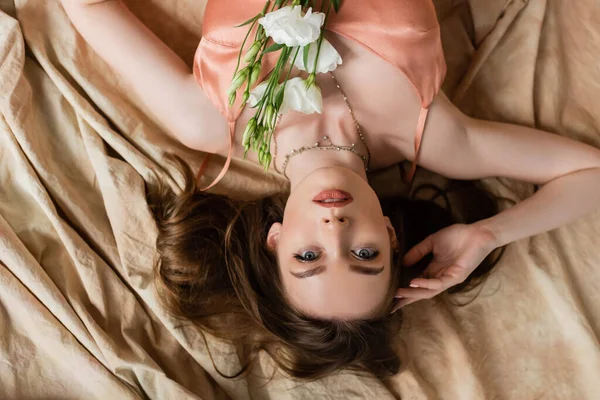 Vue du dessus de séduisante jeune femme en soie rose robe glissante couchée sur tissu de lin avec des fleurs blanches délicates et regardant la caméra sur fond beige, sensualité, vue à l'envers — Photo de stock