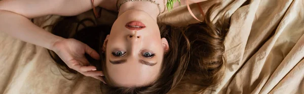 Vista dall'alto di seducente e bruna giovane donna sdraiata su tessuto di lino e guardando la fotocamera su sfondo beige, sensualità, vista a testa in giù, banner — Foto stock