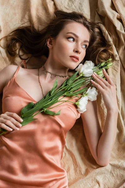 Верхний вид привлекательной молодой женщины в розовом шелковом платье скольжения лежа на льняной ткани и держа нежные белые цветы на бежевом фоне, чувственность, элегантность, эустома, глядя в сторону — стоковое фото