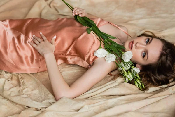 Atractiva mujer joven en vestido de seda rosa deslizamiento acostado en tela de lino y la celebración de delicadas flores blancas sobre fondo beige moteado, sensualidad, sofisticación, elegancia, eustoma, mirando a la cámara - foto de stock