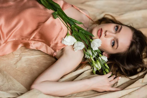 Jovem mulher de seda rosa vestido deslizante deitado sobre tecido de linho e segurando flores brancas no fundo bege, sensualidade, sofisticação, elegância, eustoma, olhando para a câmera, pose graciosa — Fotografia de Stock