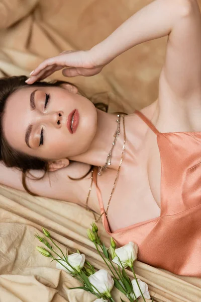 Seductive young woman with brunette hair and closed eyes lying in sexy slip dress on linen fabric near delicate white flowers, sophistication,  sensuality, elegance, eustoma — Stock Photo