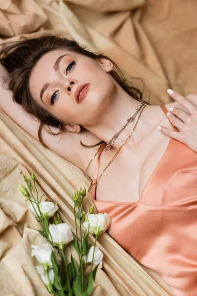 Seductive young woman with brunette hair lying in sexy slip dress on linen fabric near delicate white flowers and looking at camera, beige background, sophistication, sensuality, elegance, eustoma — Stock Photo