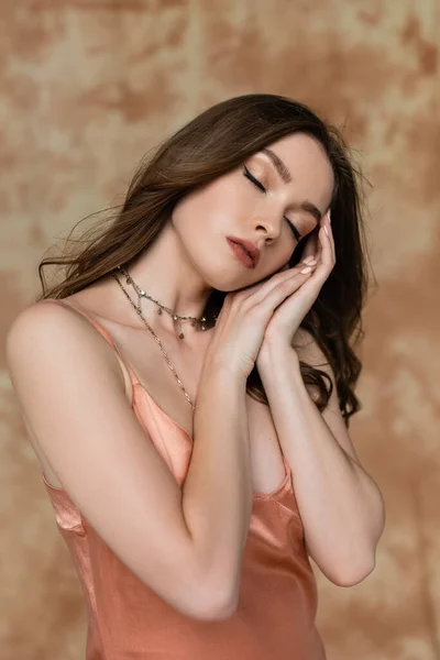 Mujer joven soñolienta con cabello moreno con sofisticado collar de oro posando con los ojos cerrados en vestido de deslizamiento rosa sobre fondo beige moteado, sensualidad, elegancia, manos cerca de la cara - foto de stock